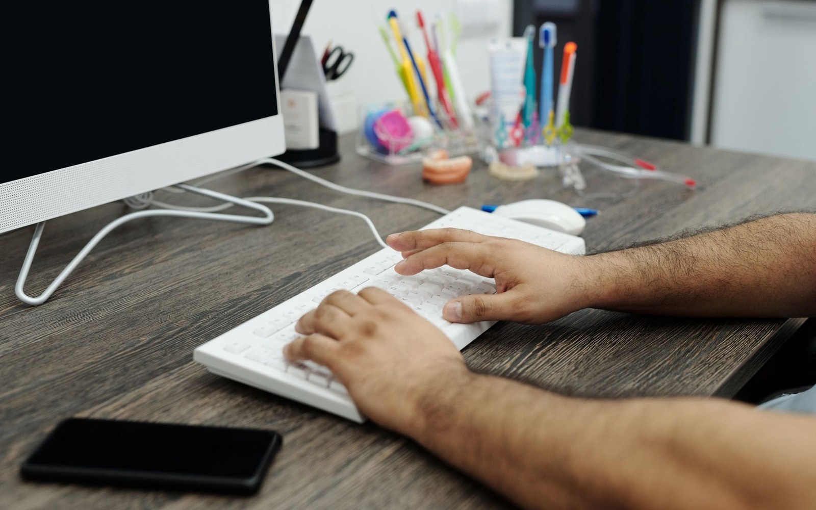 Hands typing on a white keyboard at a desk with dental tools and a monitor