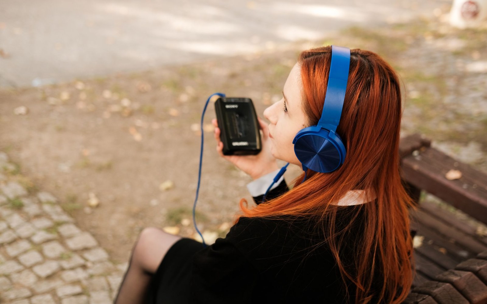 Woman with red hair wearing blue headphones holds a Sony device on a park bench