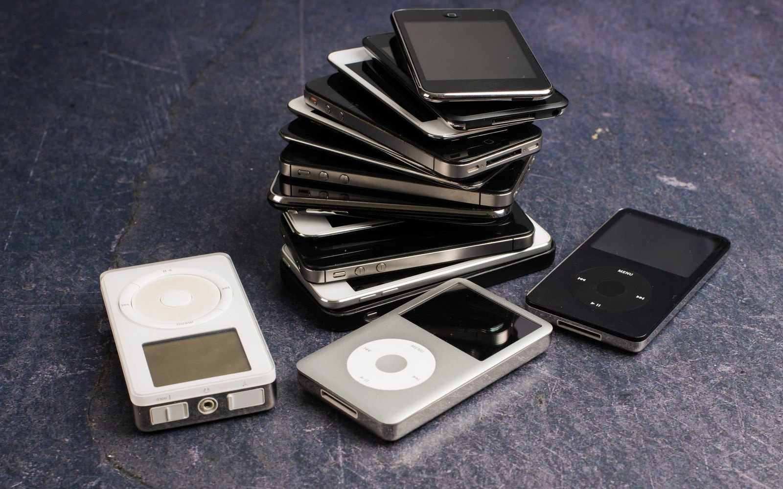 A stack of old smartphones beside several iPod media players on a dark surface